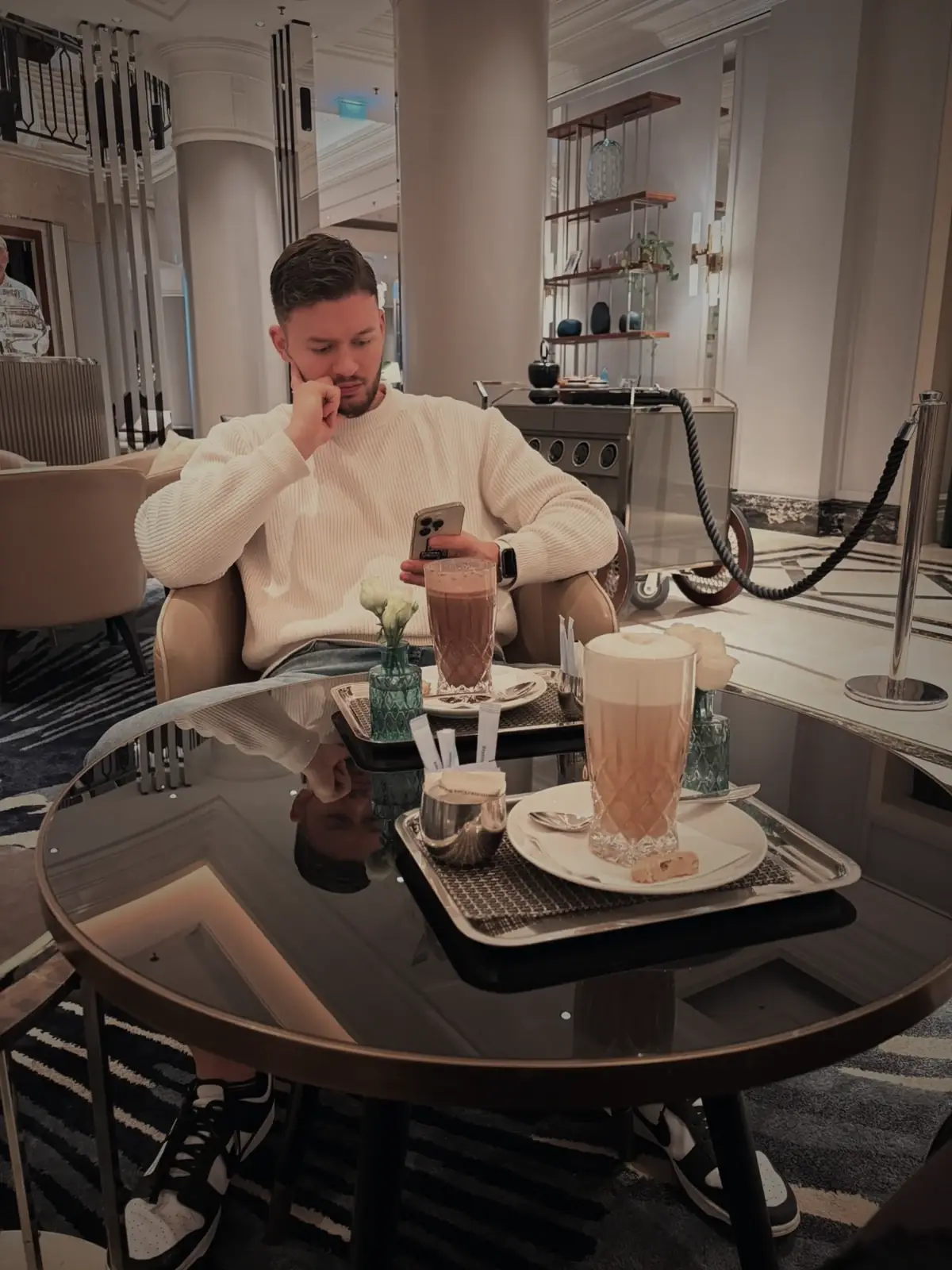 Mann mit Smartphone in einer Hotel-Lobby, vor ihm Kaffee und heiße Schokolade
