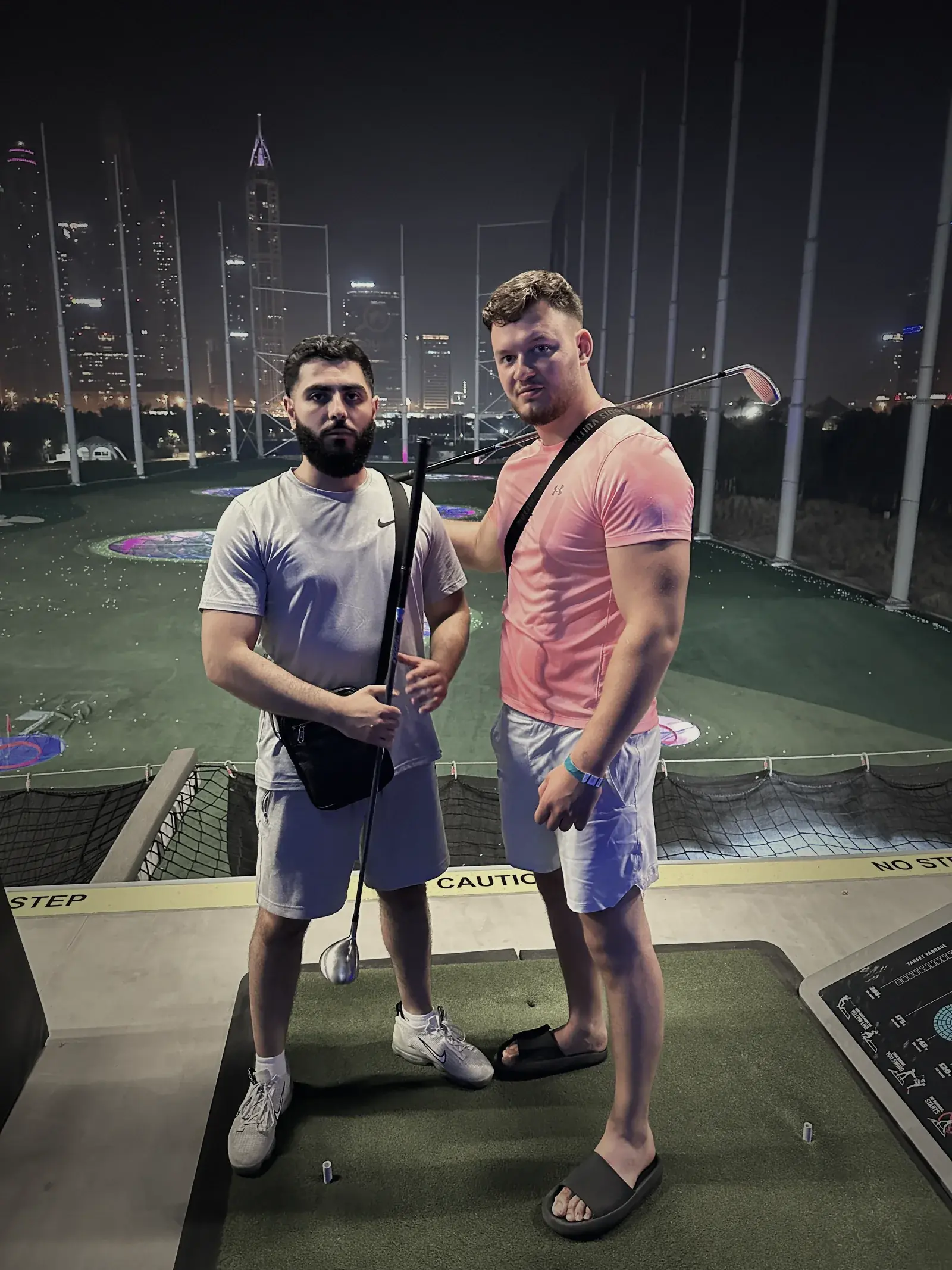 Zwei Männer mit Golfschlägern bei Nacht auf einer Driving Range vor einer beleuchteten Skyline